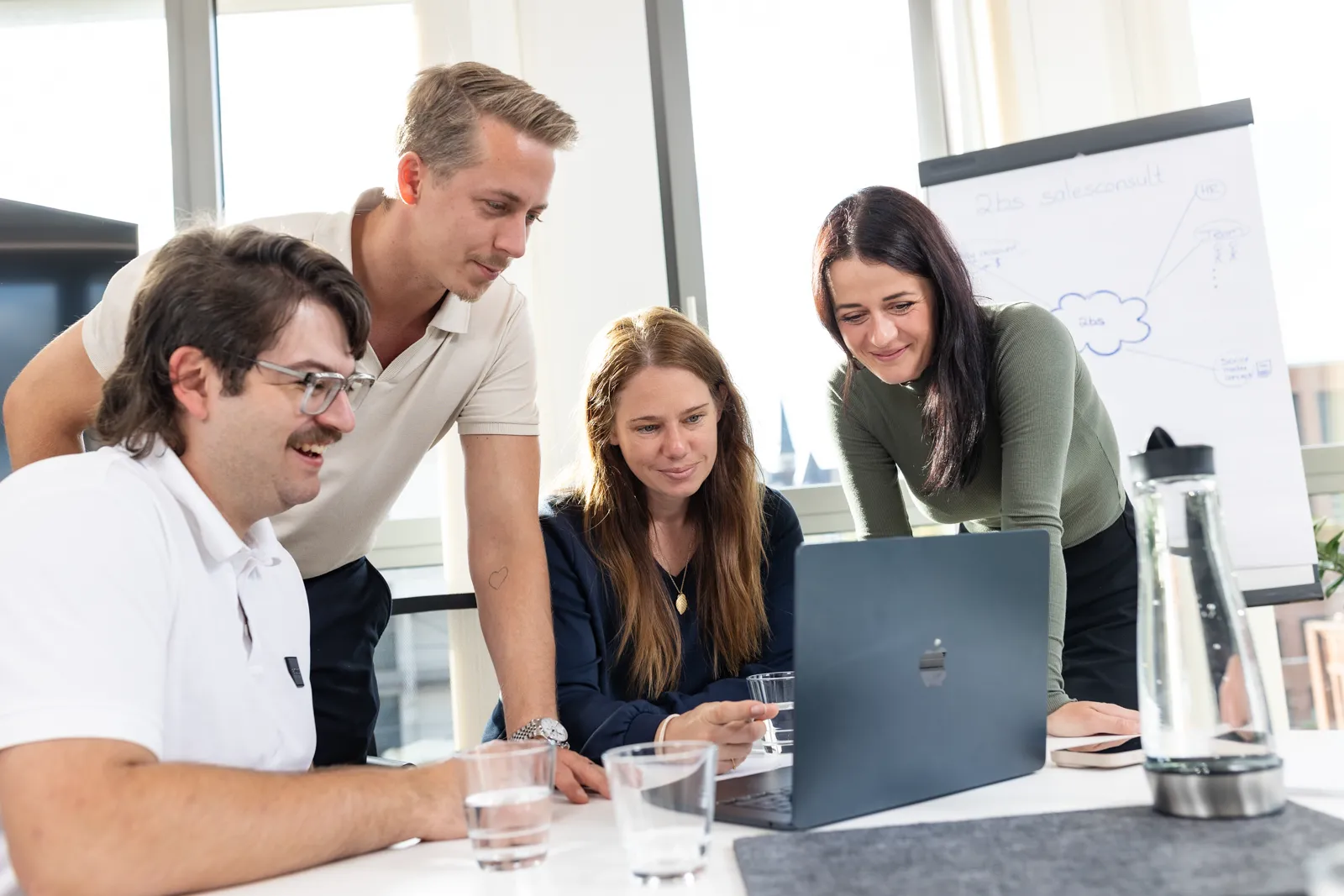 Vier Personen 2BS schauen gemeinsam lächelnd auf einen MacBook in einem hellen Büro mit Flipchart im Hintergrund. Auf dem Tisch stehen mehrere Wassergläser und eine Karaffe.