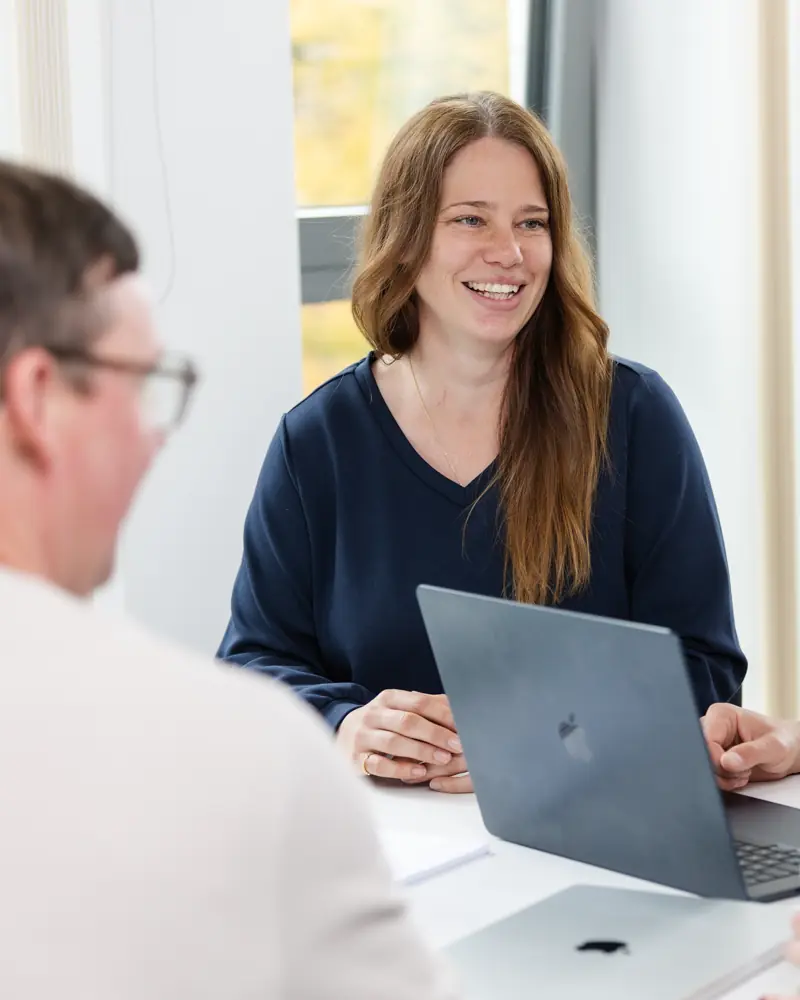 Lächelnde Frau in dunkelblauem Pullover sitzt an einem Tisch mit offenem Laptop vor sich, zwei Personen im Vordergrund unscharf.