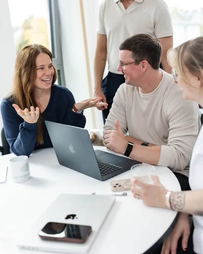 Drei lächelnde Personen sitzen an einem runden Tisch mit einem Laptop und diskutieren lebhaft.