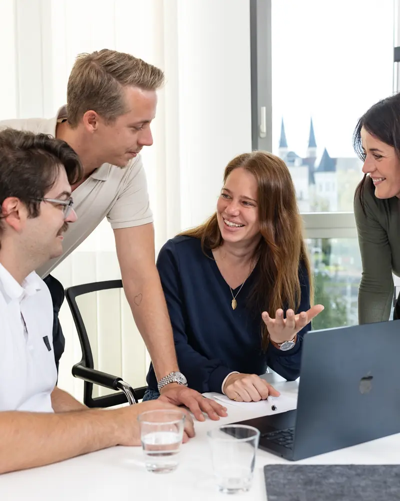 Vier junge Erwachsene diskutieren und lächeln gemeinsam um einen Tisch mit Laptop in einem hellen Büroraum.
