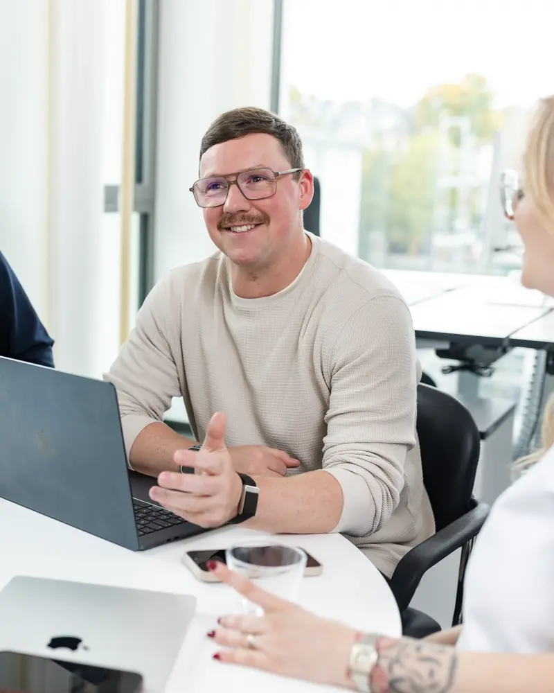 Ein lächelnder Mann mit Brille und hellbeigem Pullover sitzt an einem Tisch und spricht mit einer Frau, im Hintergrund ein Laptop.