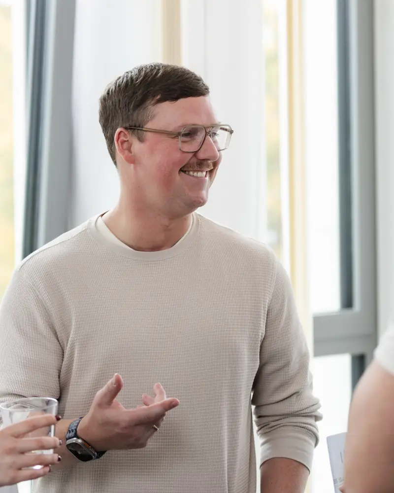 Ein lächelnder Mann mit Brille und Pullover steht in einem hellen Raum und gestikuliert mit der Hand.