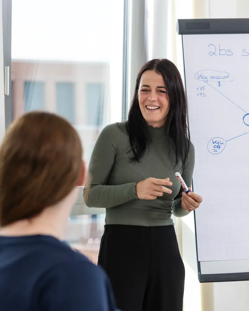 Frau lächelt und hält einen Marker vor einem Flipchart mit handschriftlichen Notizen, während sie mit einem anderen Menschen spricht.
