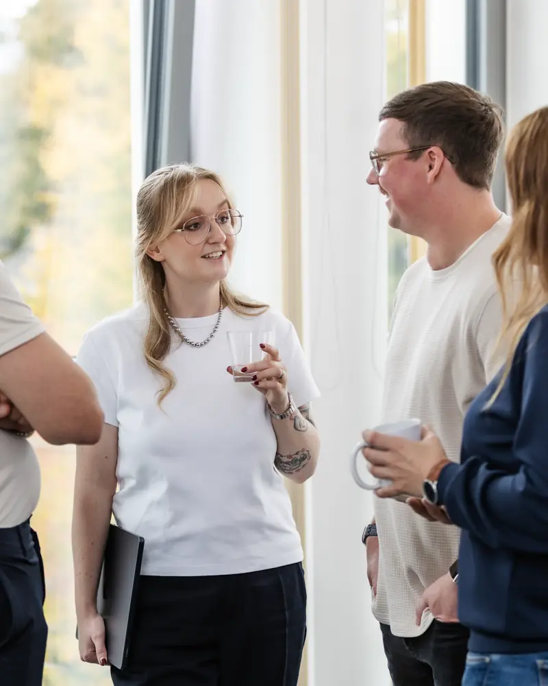 Vier junge Berufstätige unterhalten sich entspannt mit Getränken in der Hand in einem hellen Raum mit großen Fenstern.