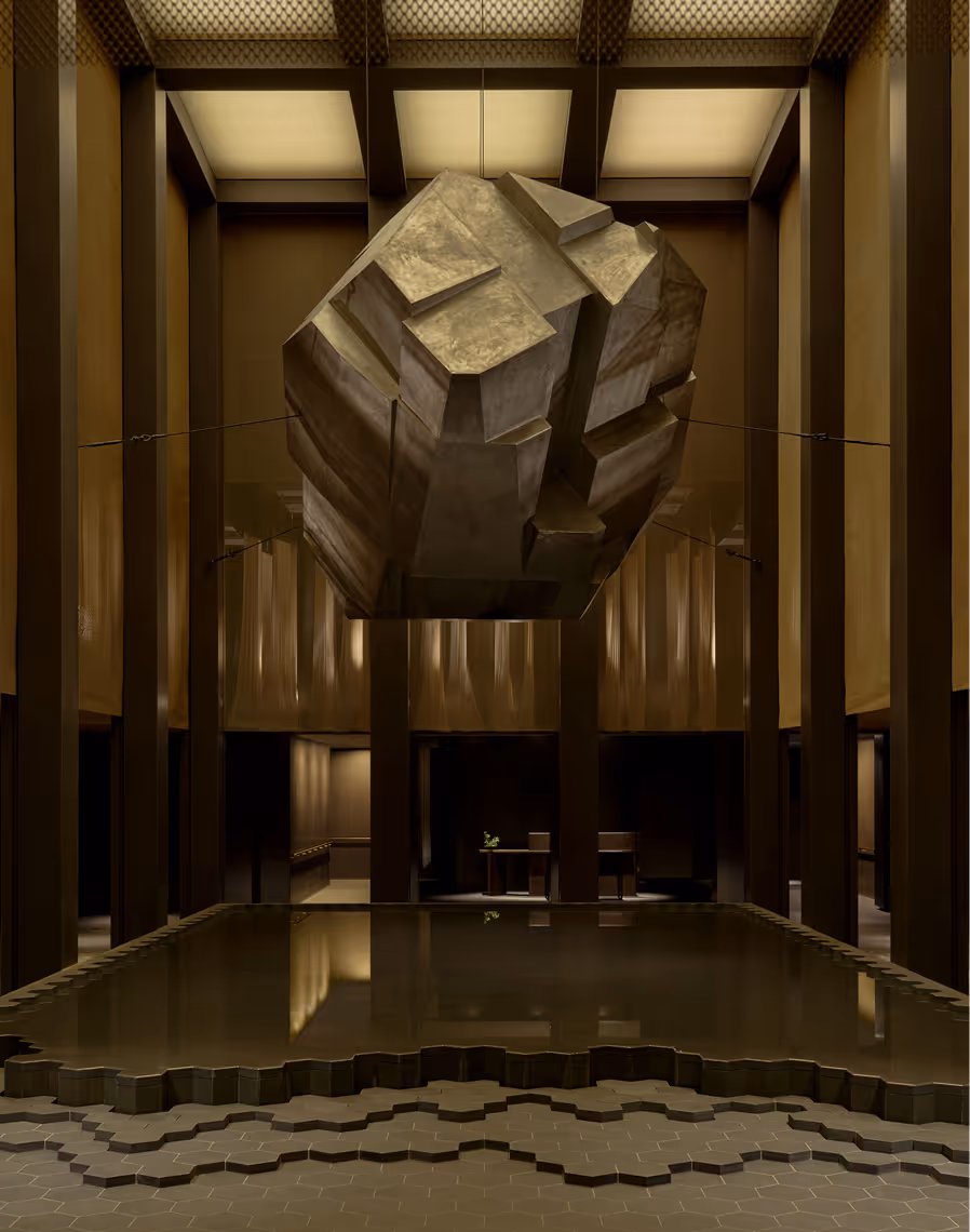 Large geometric stone sculpture suspended above a reflective pool in a dimly lit, modern interior space.
