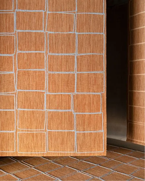 Close-up of orange-brown rugs with a grid pattern of white stitched squares, displayed indoors on floor and hanging.