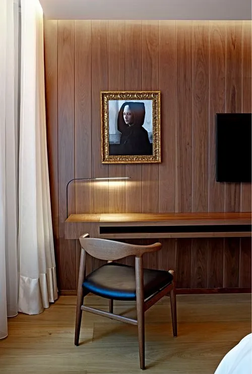 Modern wooden chair with black seat cushion in front of a wall-mounted wooden desk under a gold-framed portrait on a wood-paneled wall.