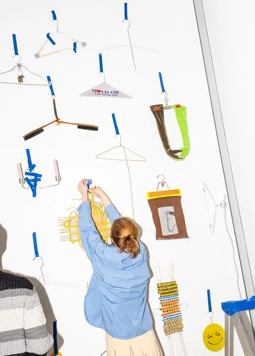 Person in a blue shirt attaching a hanger to a white wall decorated with various colorful and uniquely shaped hangers and objects.