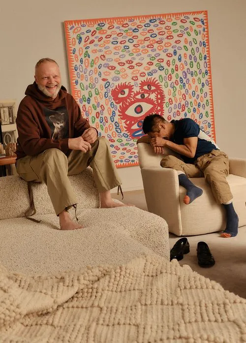 Two men sitting in a room with a colorful abstract artwork behind them; one man is barefoot and smiling, the other leans forward resting his head on his arm.
