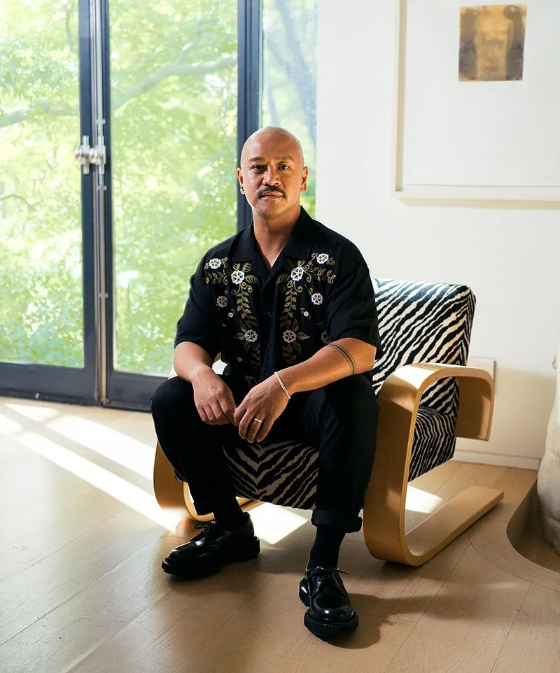 A bald man with a mustache wearing a black floral shirt and black pants sitting on a zebra-patterned chair in a bright room with large windows.