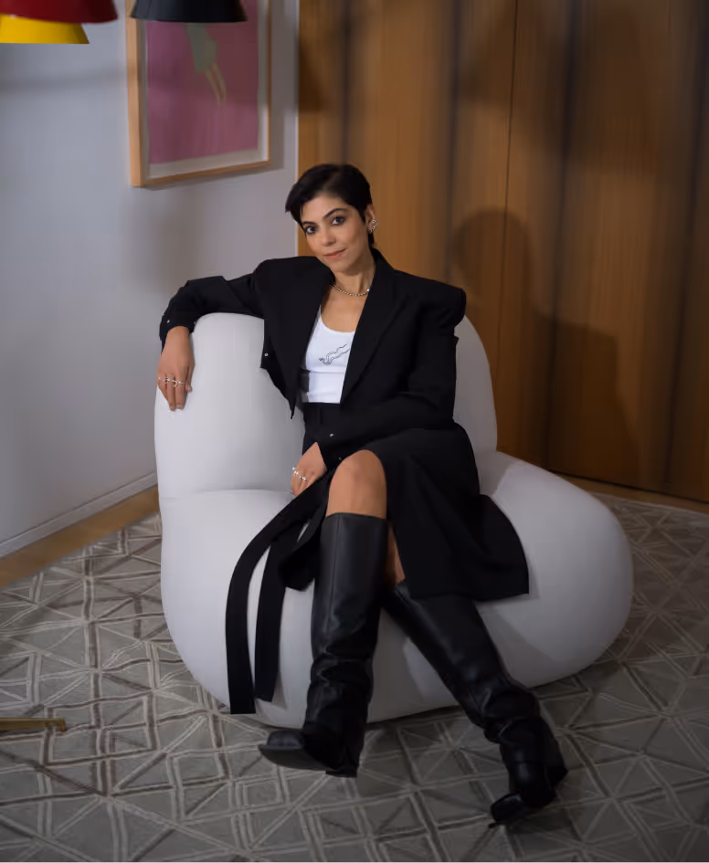 Woman with short dark hair wearing a black blazer, black skirt, white top, and black knee-high boots sitting on a modern white chair.