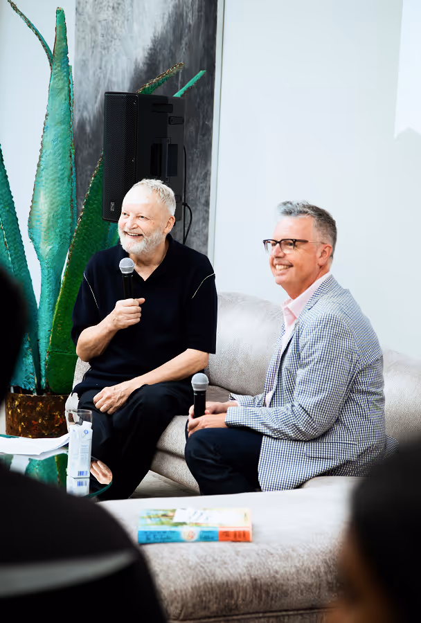 Two men sitting on a couch holding microphones, engaged in a discussion with a large green plant and speaker behind them.
