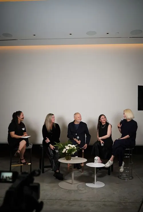 Five people sitting on chairs and stools on a stage, engaged in a panel discussion with microphones and a flower arrangement on tables in front of them.