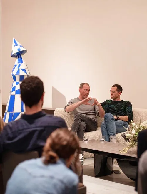Two men seated on a couch engaged in conversation during a group discussion with several people facing them.
