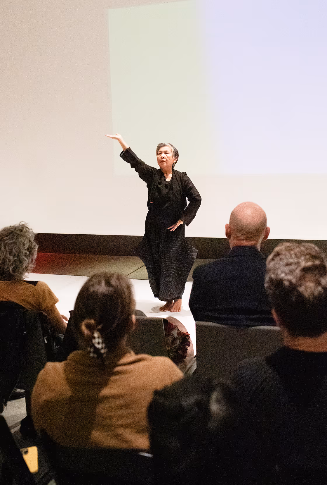 Barefoot woman in black clothing performing a dance or pose on stage in front of a seated audience.
