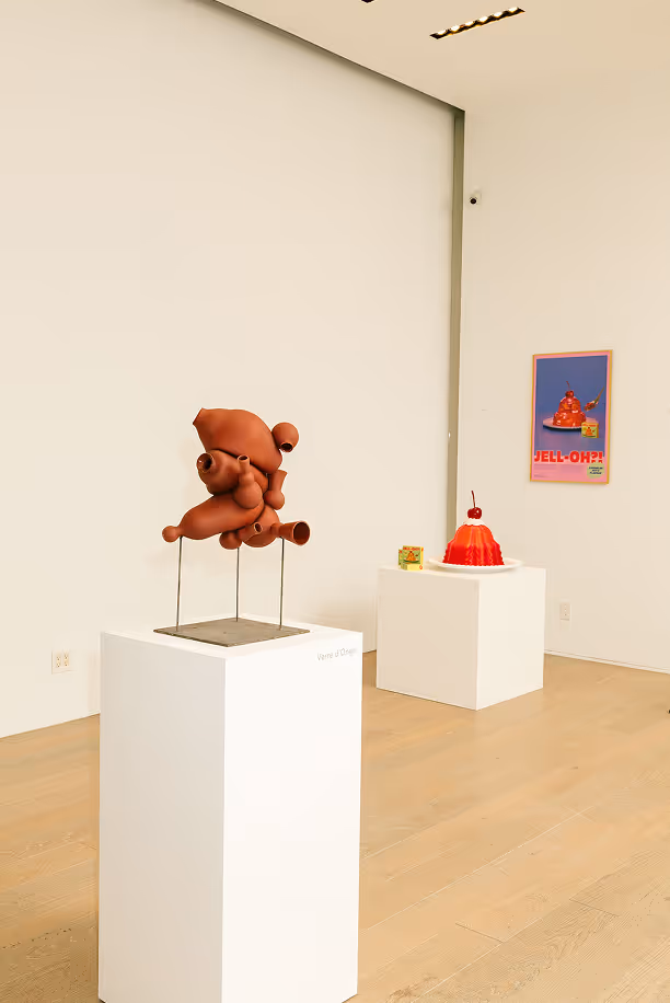 Abstract brown ceramic sculpture on a pedestal in a minimalist gallery space, with a red Jell-O dessert model and poster in the background.