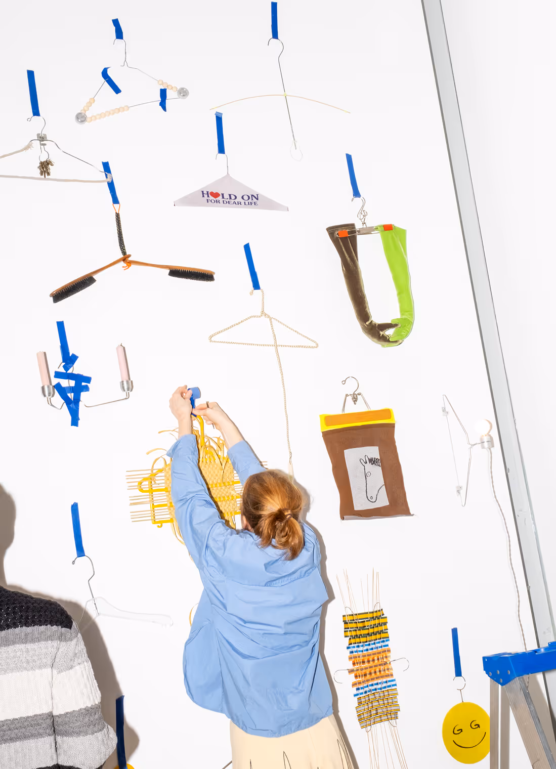 Person with brown hair tied back wearing a blue shirt is hanging artistic and colorful hangers taped with blue tape on a white wall.