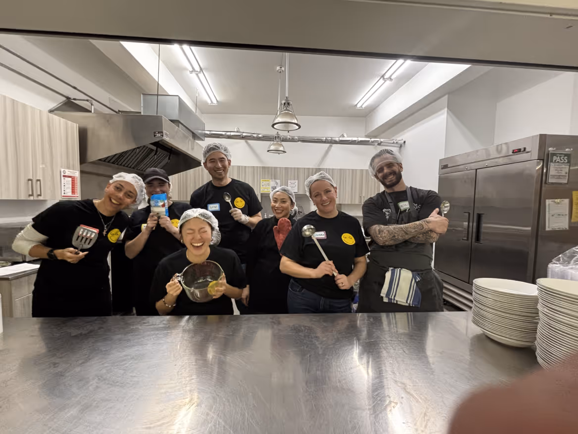 Group of seven people wearing hairnets and black shirts smiling in a commercial kitchen, some holding cooking utensils and a mixing bowl.