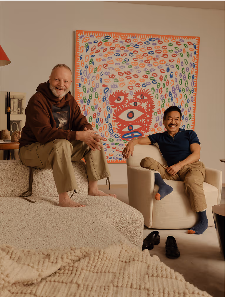 Two men smiling and relaxing in a living room with a colorful abstract eye painting on the wall behind them.