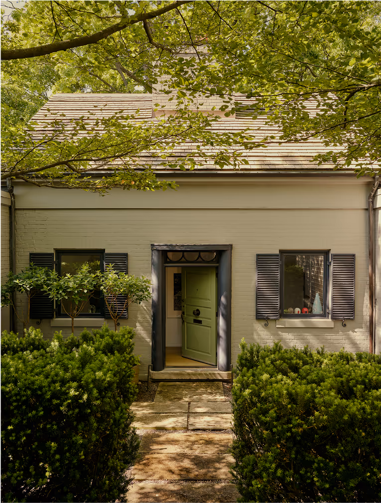 Front view of a beige bungalow with an open green door, black window shutters, surrounded by green bushes and tree branches.