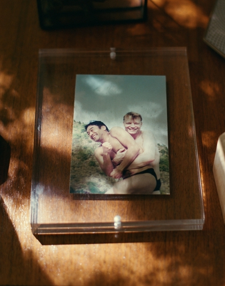 Photo of two men in swimwear hugging and smiling outdoors, mounted in a clear display case on a wooden surface.