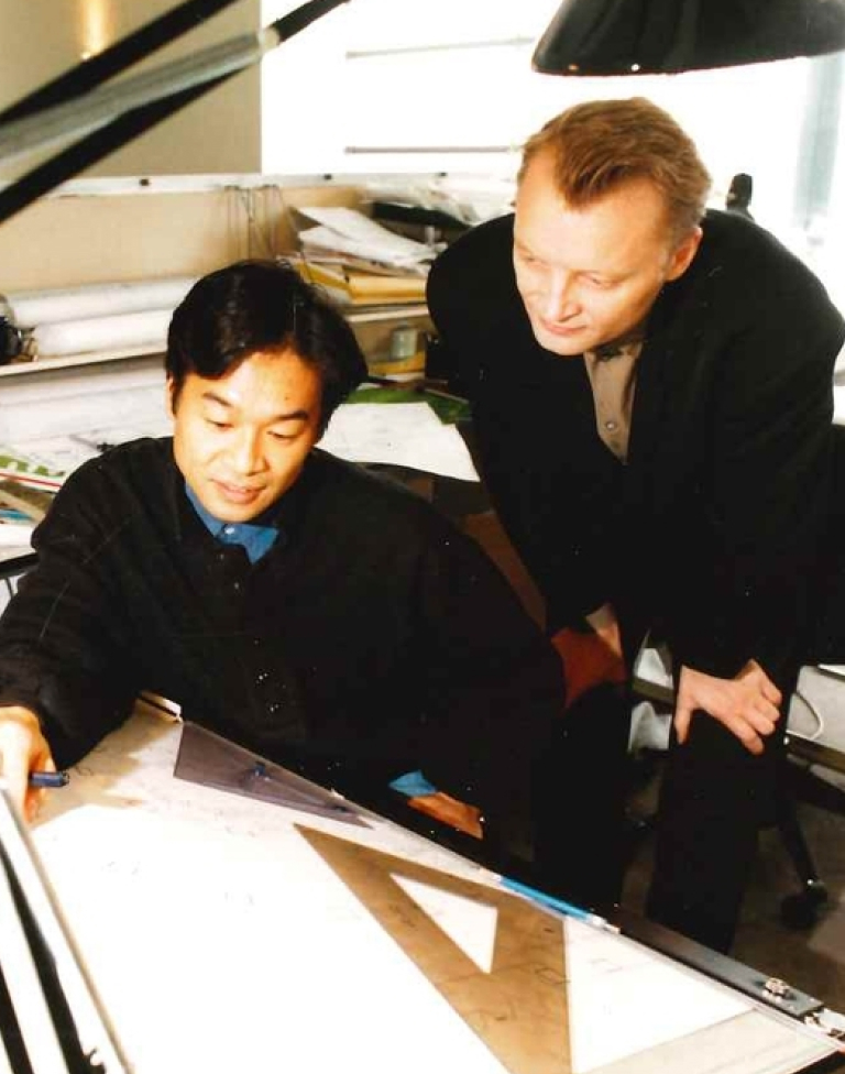 Two men working together at a drafting table with architectural tools and drawings.