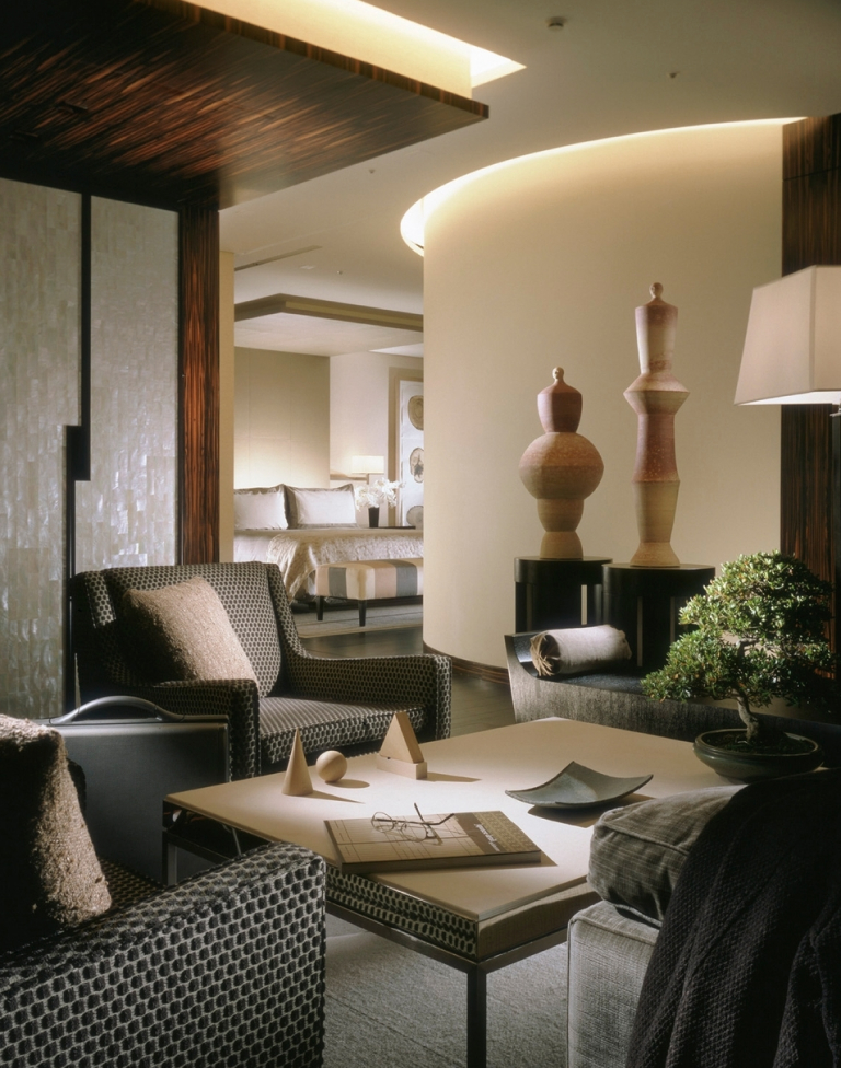 Modern living room with patterned armchairs, a coffee table with geometric sculptures and a book, a bonsai plant, and soft warm lighting, adjacent to a bedroom.