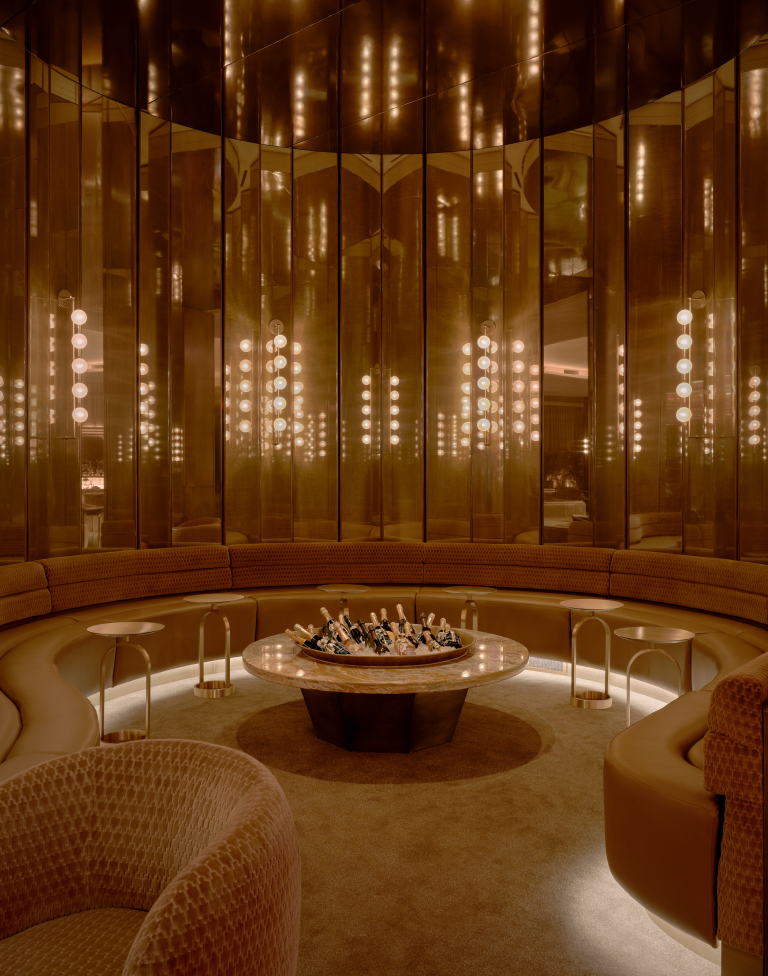 Luxurious curved lounge area with leather seating surrounding a round marble table holding multiple bottles of champagne, reflected in tall mirrored panels with warm, ambient lighting.