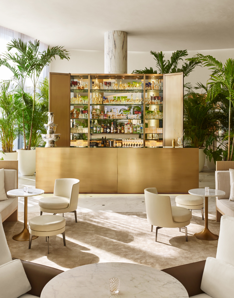 Elegant bar area with a brass counter, glass shelves stocked with bottles and glassware, surrounded by beige chairs and green potted plants.