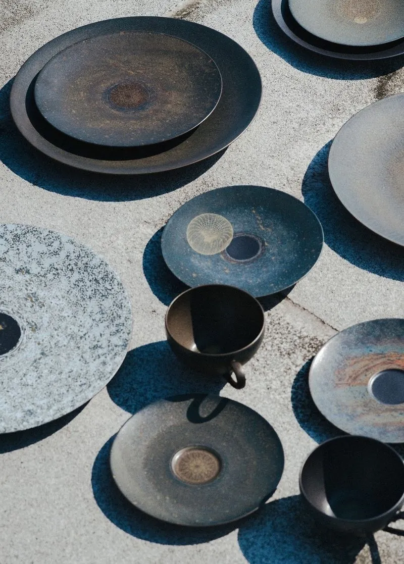 Various dark ceramic plates and cups arranged on a textured concrete surface casting shadows.