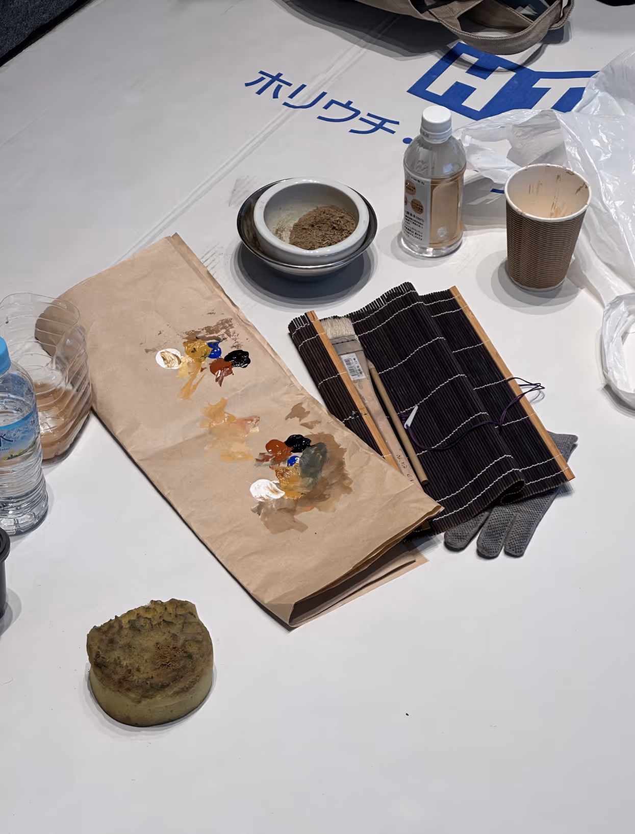 Table with painting supplies including brushes, a paper palette with paint, a sponge, a bowl with powder, and a plastic bottle on a white surface with Japanese text and a blue logo.