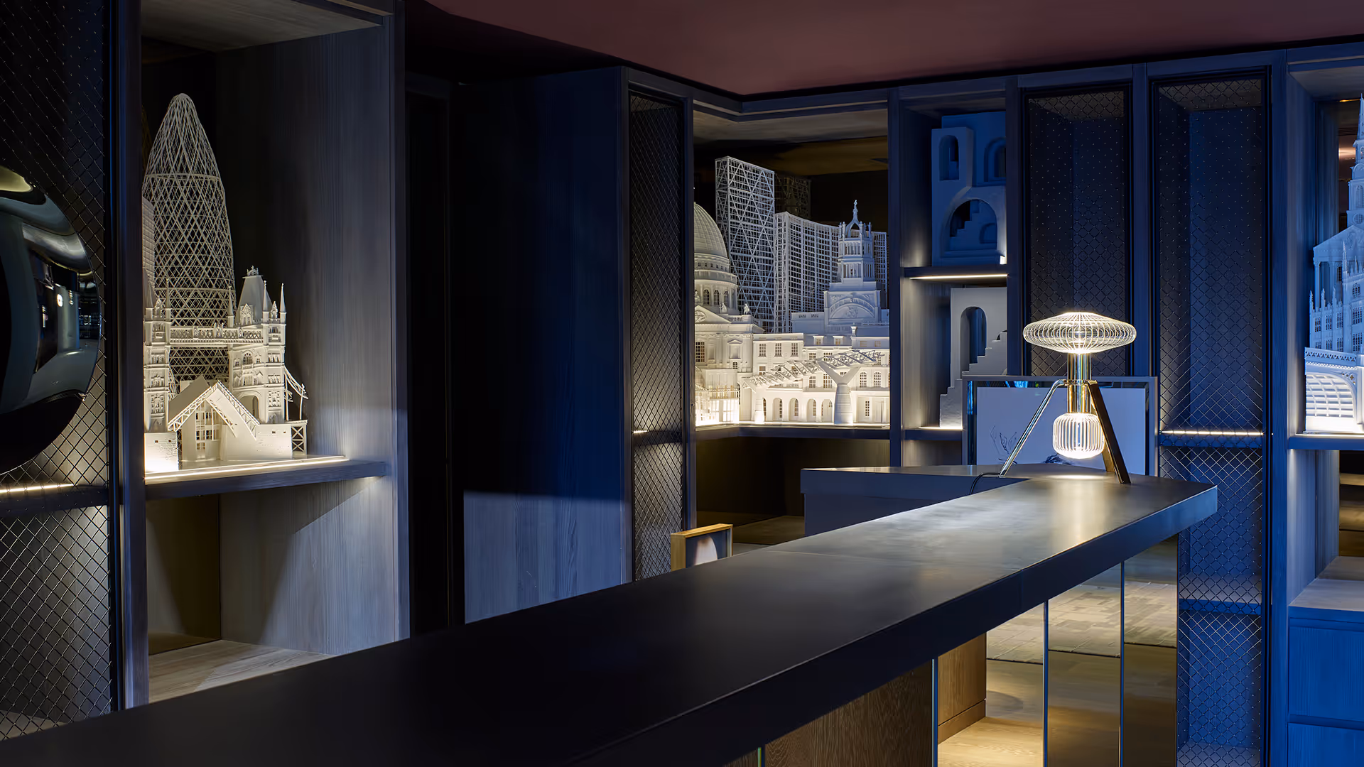 Modern interior with dark wood shelves displaying illuminated white architectural scale models of London landmarks and a contemporary wireframe table lamp on a sleek counter.