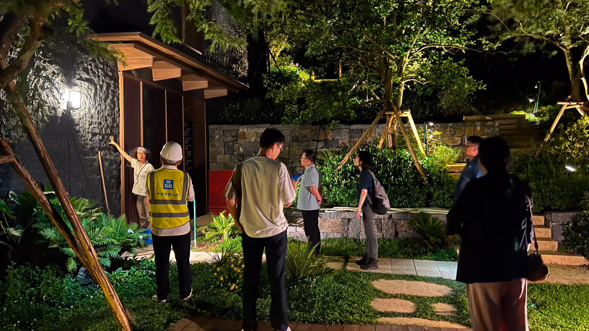 Group of people gathered outside a house at night, with one person in a safety vest and helmet listening to a woman speaking near the door.