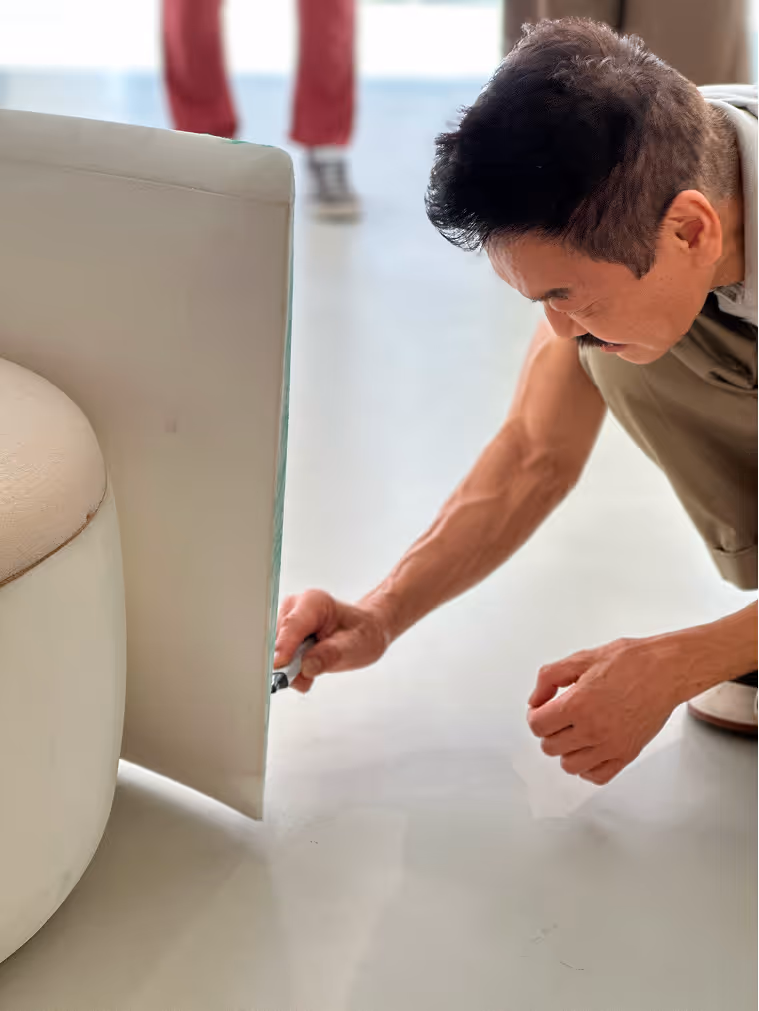 Man inspecting or fixing the bottom side of a light-colored sofa while crouching on the floor.