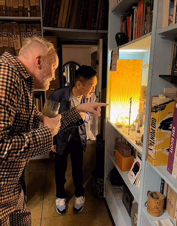 Two men closely examining a small illuminated display shelf filled with various objects and books in a cozy, dimly lit room.