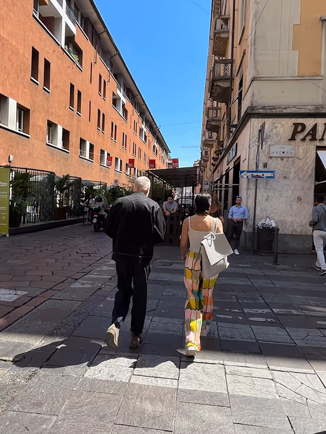 Two people walking down a narrow urban street between tall buildings on a sunny day.