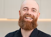 Smiling man with dark hair and light skin standing outdoors in front of green foliage.