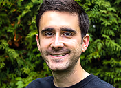 Smiling man with short dark hair and a trimmed beard standing outdoors with green foliage in the background.