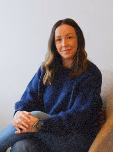 Woman with wavy hair wearing a dark blue sweater sitting with folded hands on a chair against a light background.