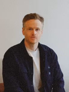 Portrait of a man with short light brown hair wearing a dark jacket over a white shirt against a plain light background.