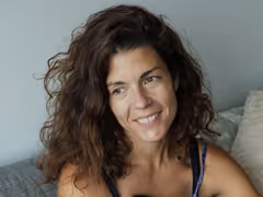 Smiling woman with curly brown hair sitting on a couch against a light blue wall.