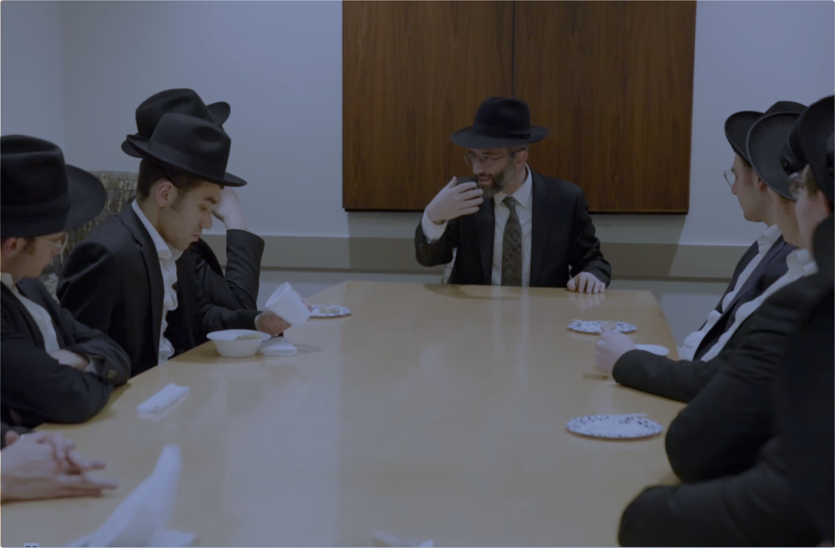 Group of men wearing black hats and suits seated around a conference table, one man at the head gesturing while speaking.