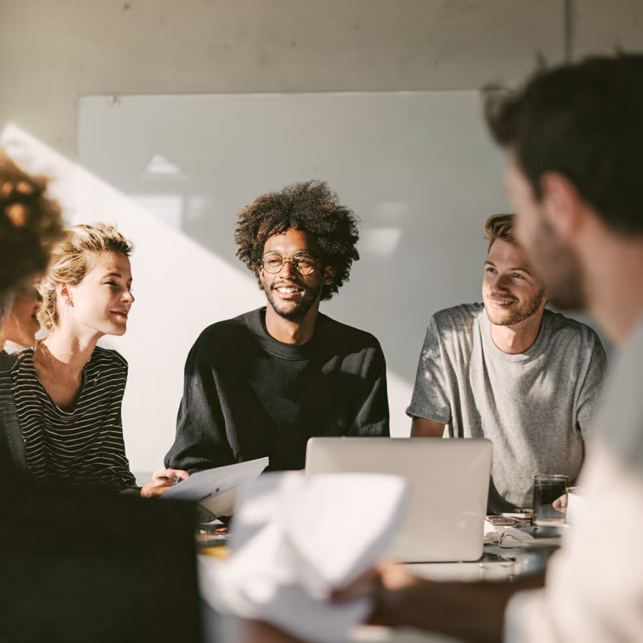 Gruppe junger Erwachsener, die in einem hellen Raum an einem Tisch mit Laptop und Papieren zusammen an einem Projekt arbeiten.