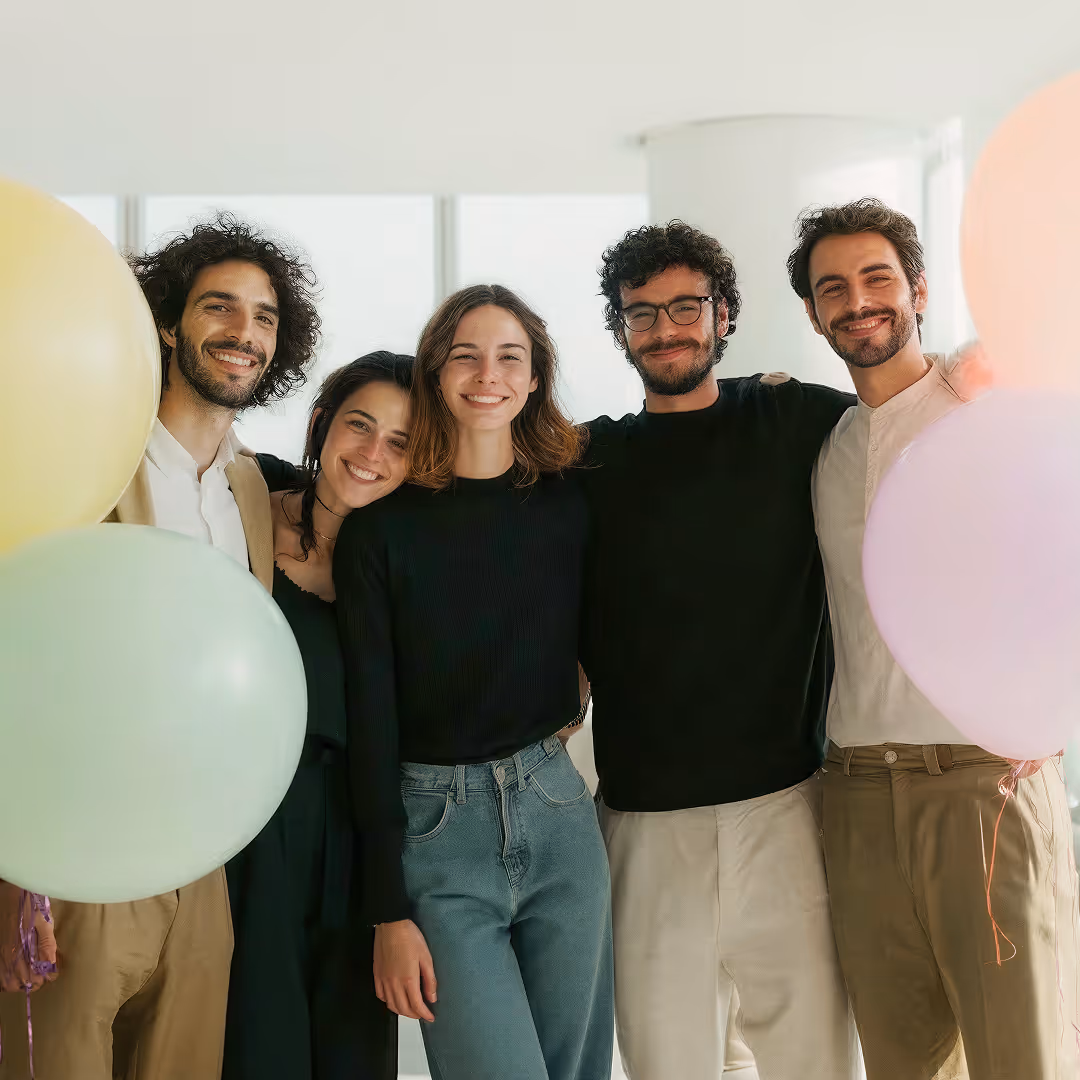 Fünf lächelnde junge Erwachsene stehen nah beieinander, umgeben von bunten Luftballons in einem hellen Raum.