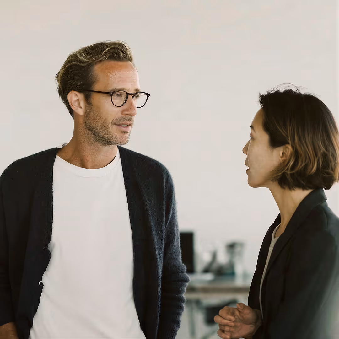 Zwei Erwachsene, ein Mann mit Brille und ein Frau, unterhalten sich in einem hellen Büro.