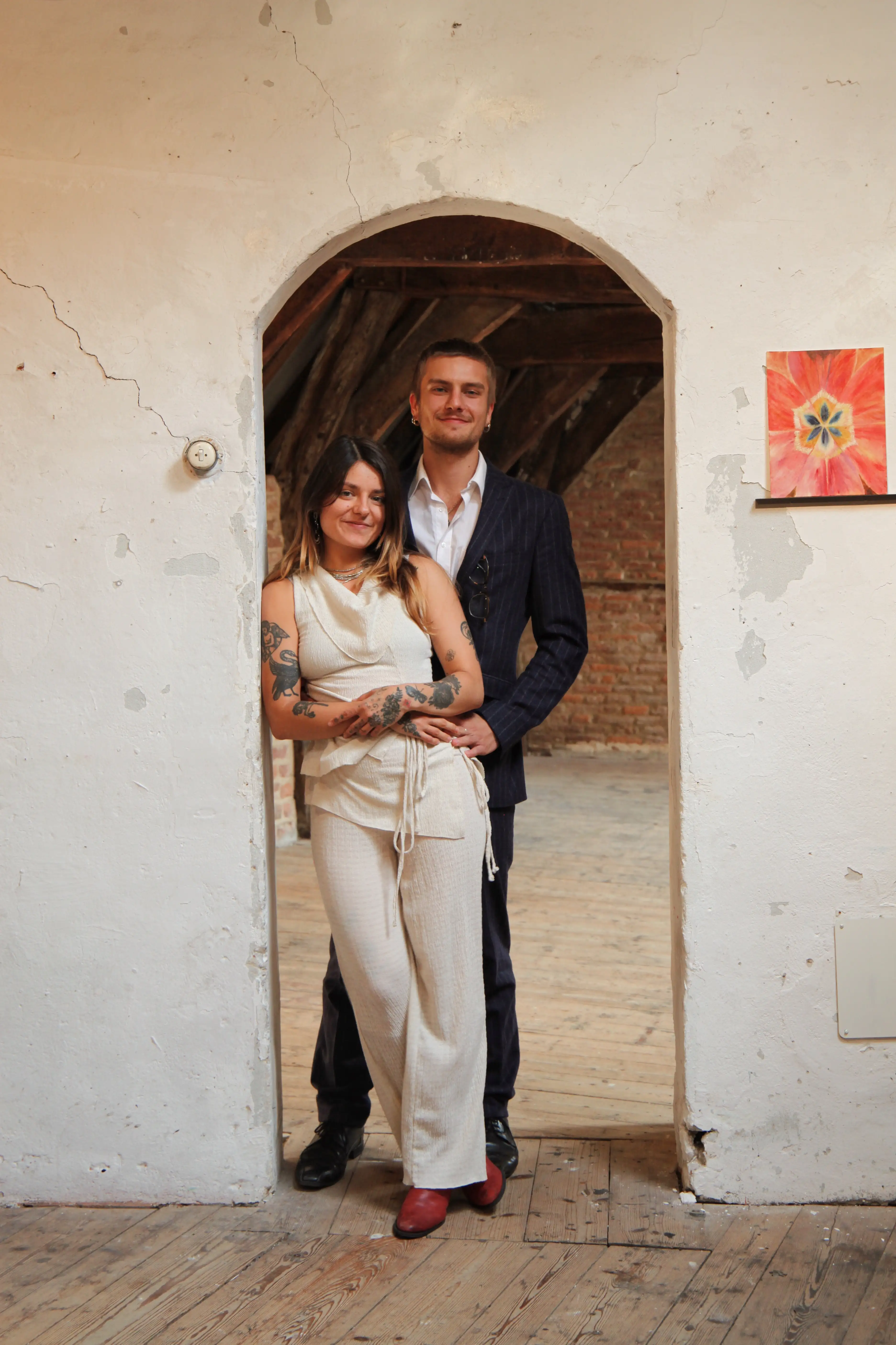 A smiling couple stands in a rustic arched doorway; the woman wears white pants with a sleeveless top and red shoes, and the man wears a dark suit with a white shirt