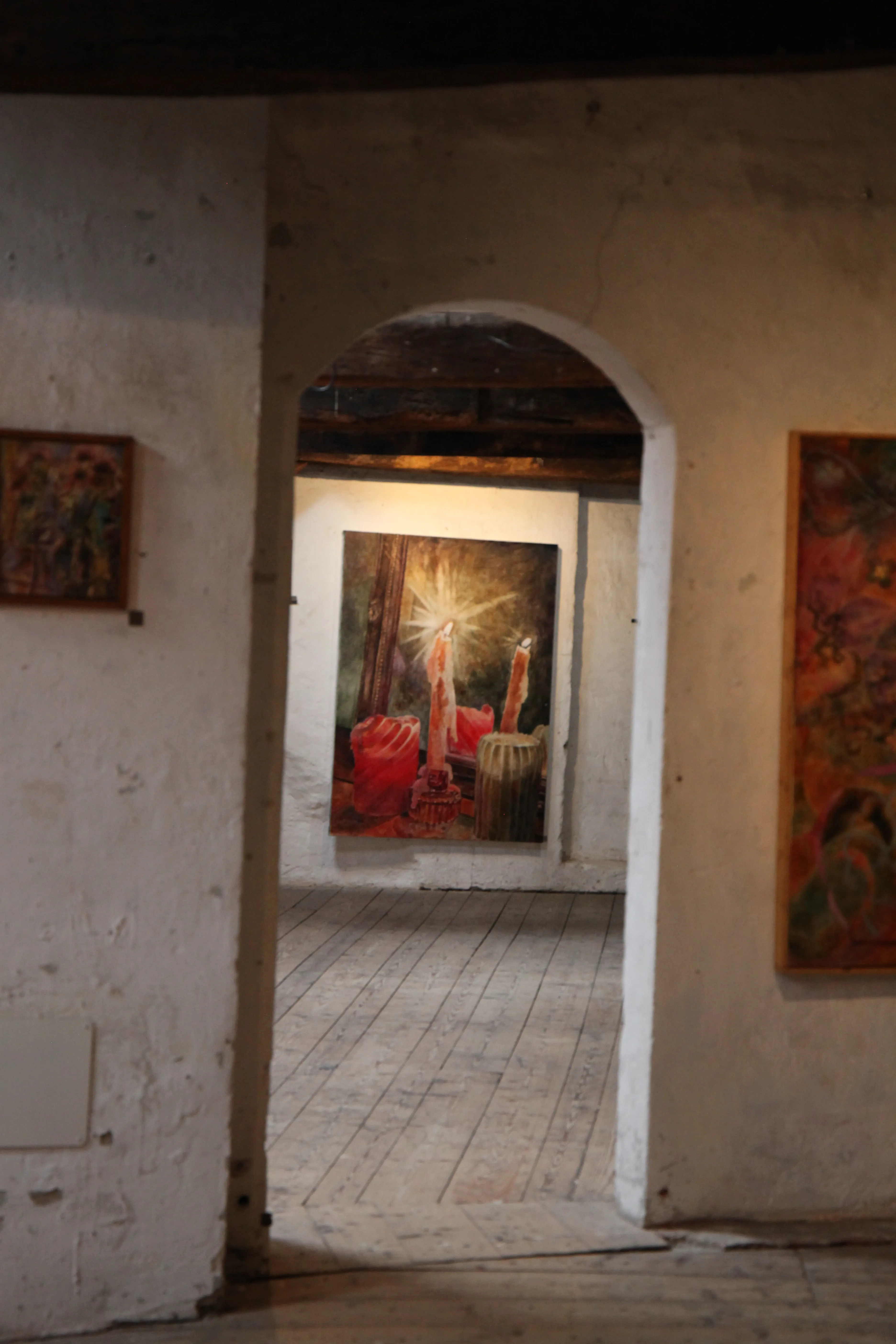 View through an archway into a room with wooden floors and a painting of three lit candles on the far wall.