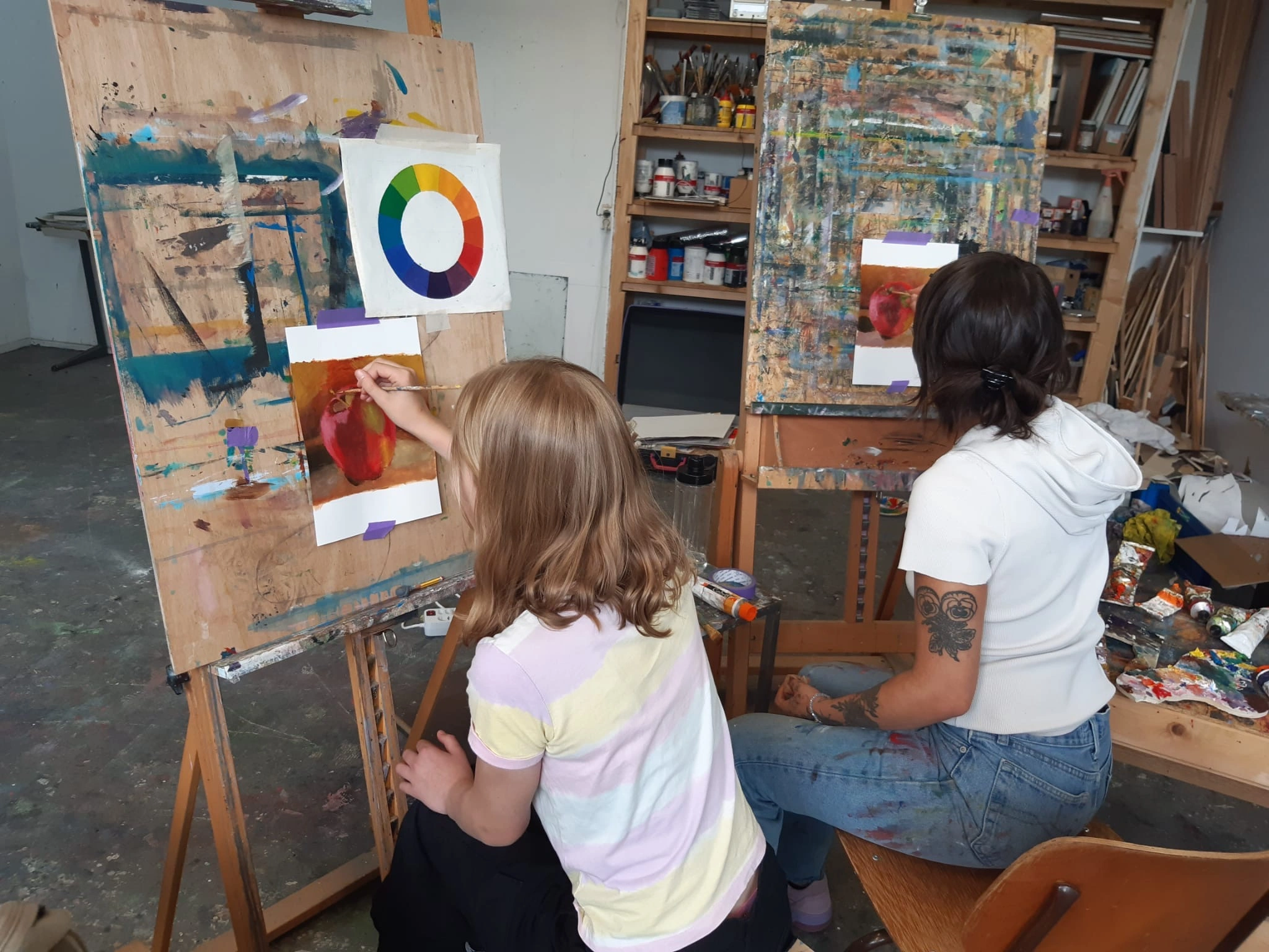 Two people sitting on stools painting apples on canvases attached to easels in an art studio with painting supplies and color wheel on one easel