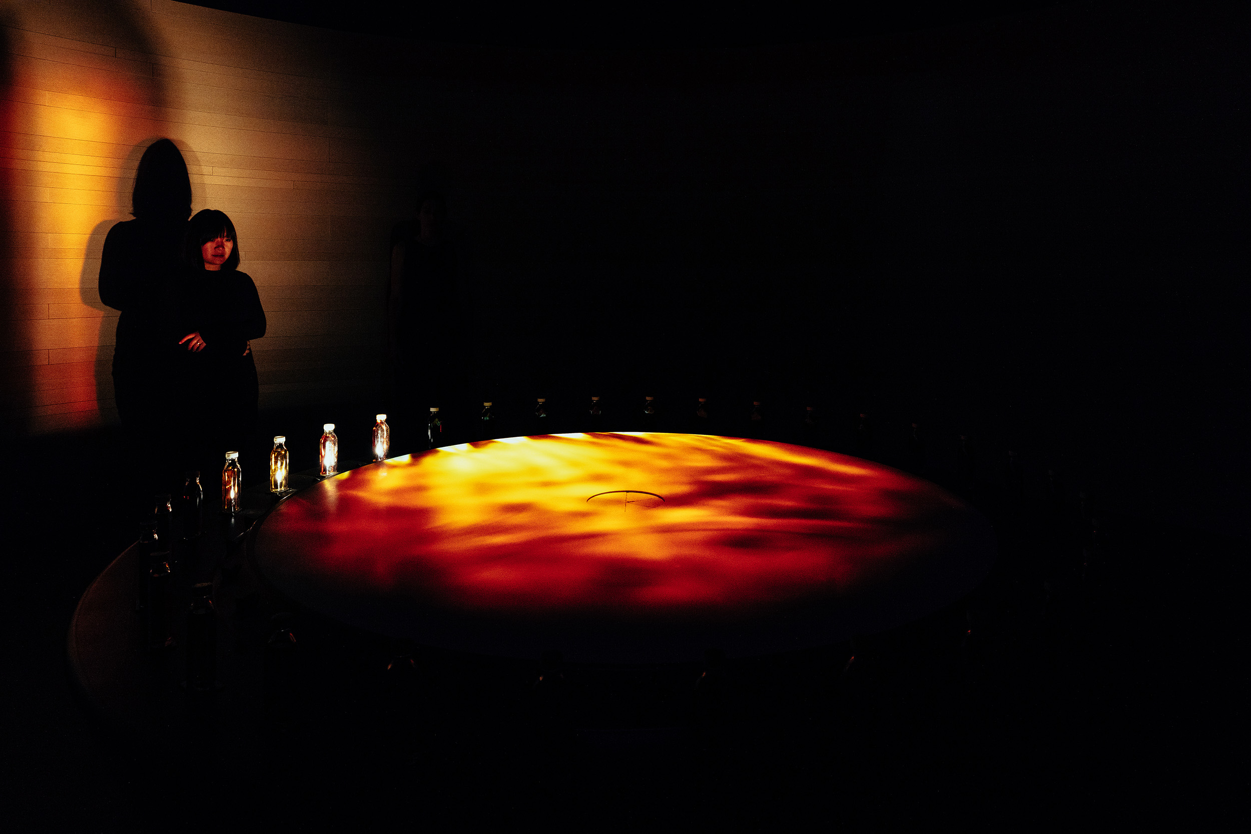 Silhouetted figure standing before glowing red-orange surface with illuminated bottles.