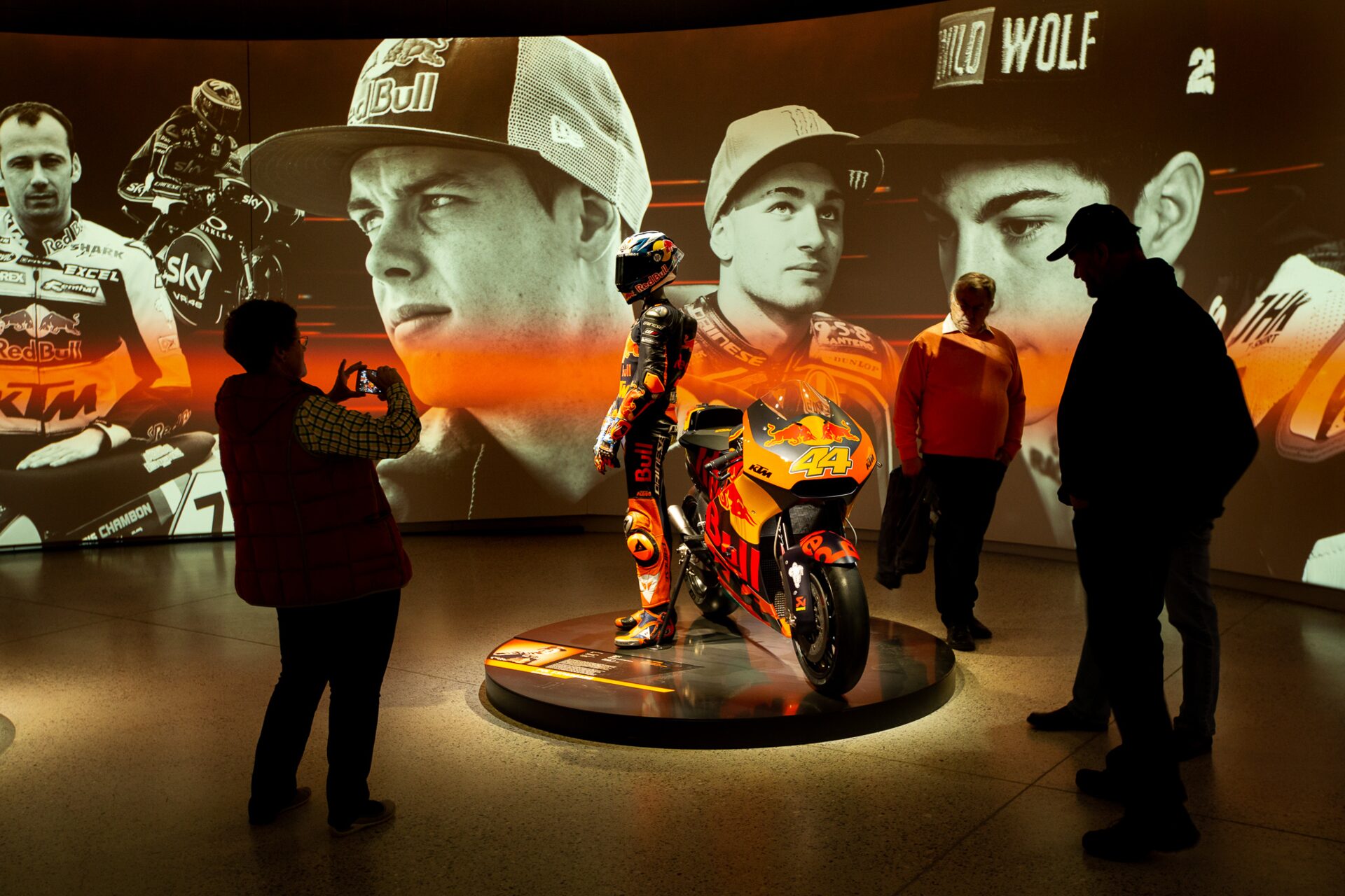 Visitors viewing KTM motorcycle exhibition with large display screens showing racing drivers and motorcycles